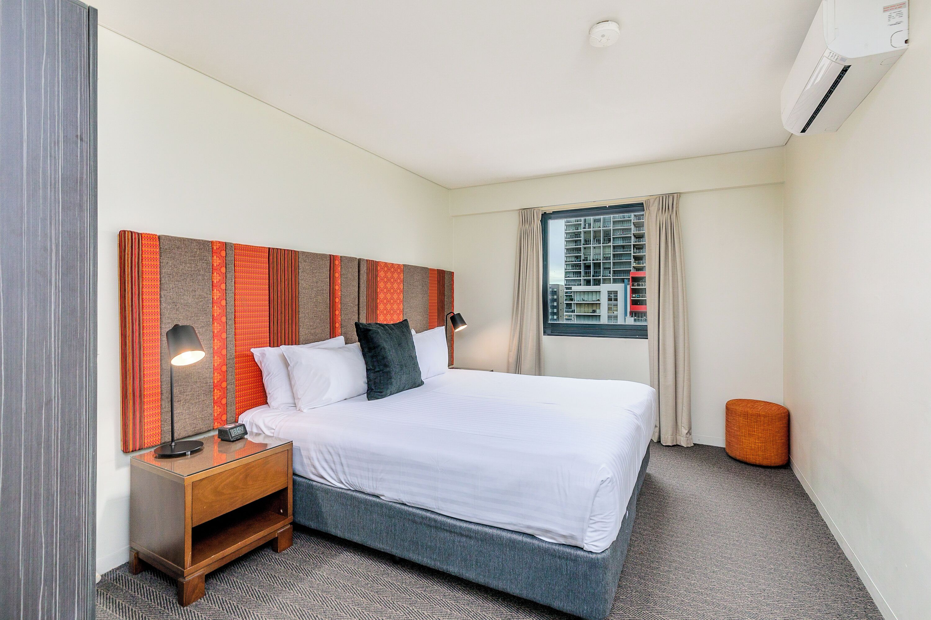 A hotel room with a bed, a nightstand, a lamp, a window with curtains, and an air conditioning unit.