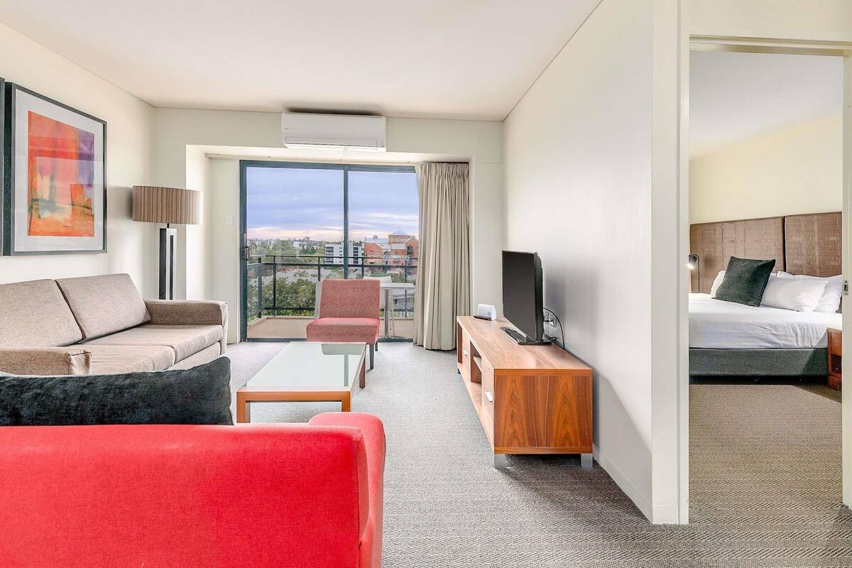 A hotel room with a sofa, a red chair, a TV, a bed, and a balcony with a city view.