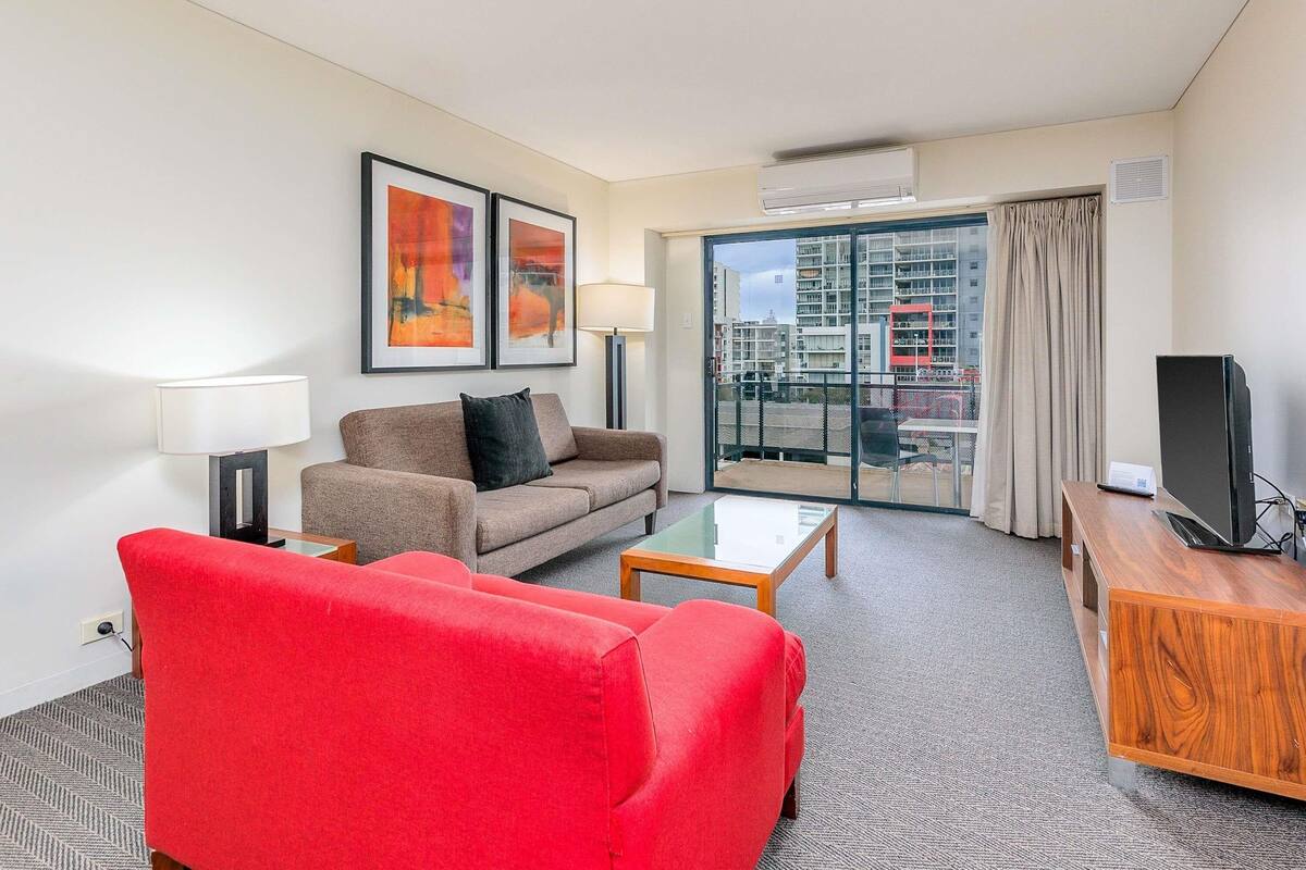 A hotel room with a red sofa, a brown sofa, a glass coffee table, a TV on a wooden stand, and a balcony with a city view.