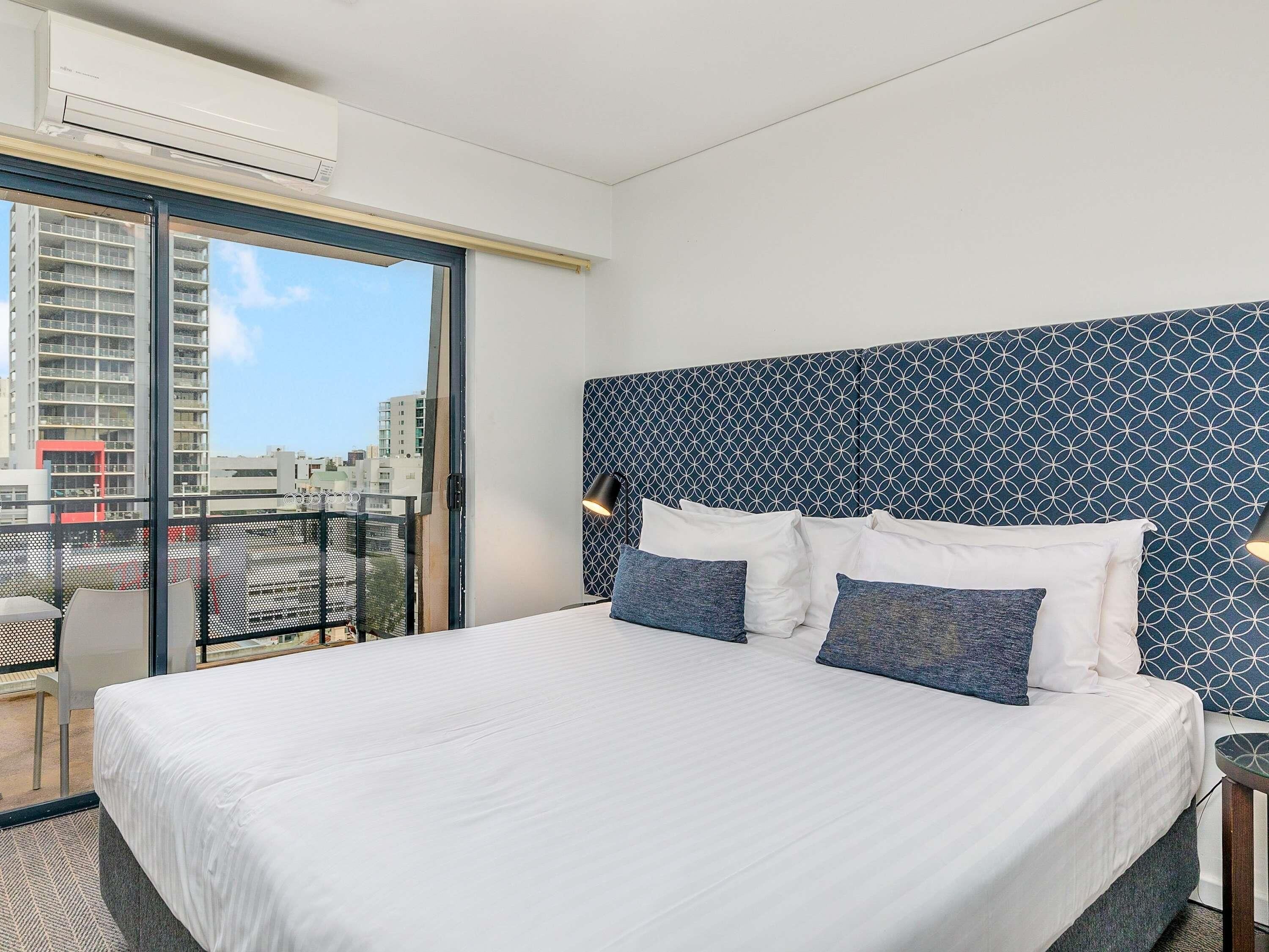 Ein Hotelzimmer mit einem großen Bett, einem gemusterten Kopfteil, einem Balkon mit Blick auf Gebäude und einer Klimaanlage.