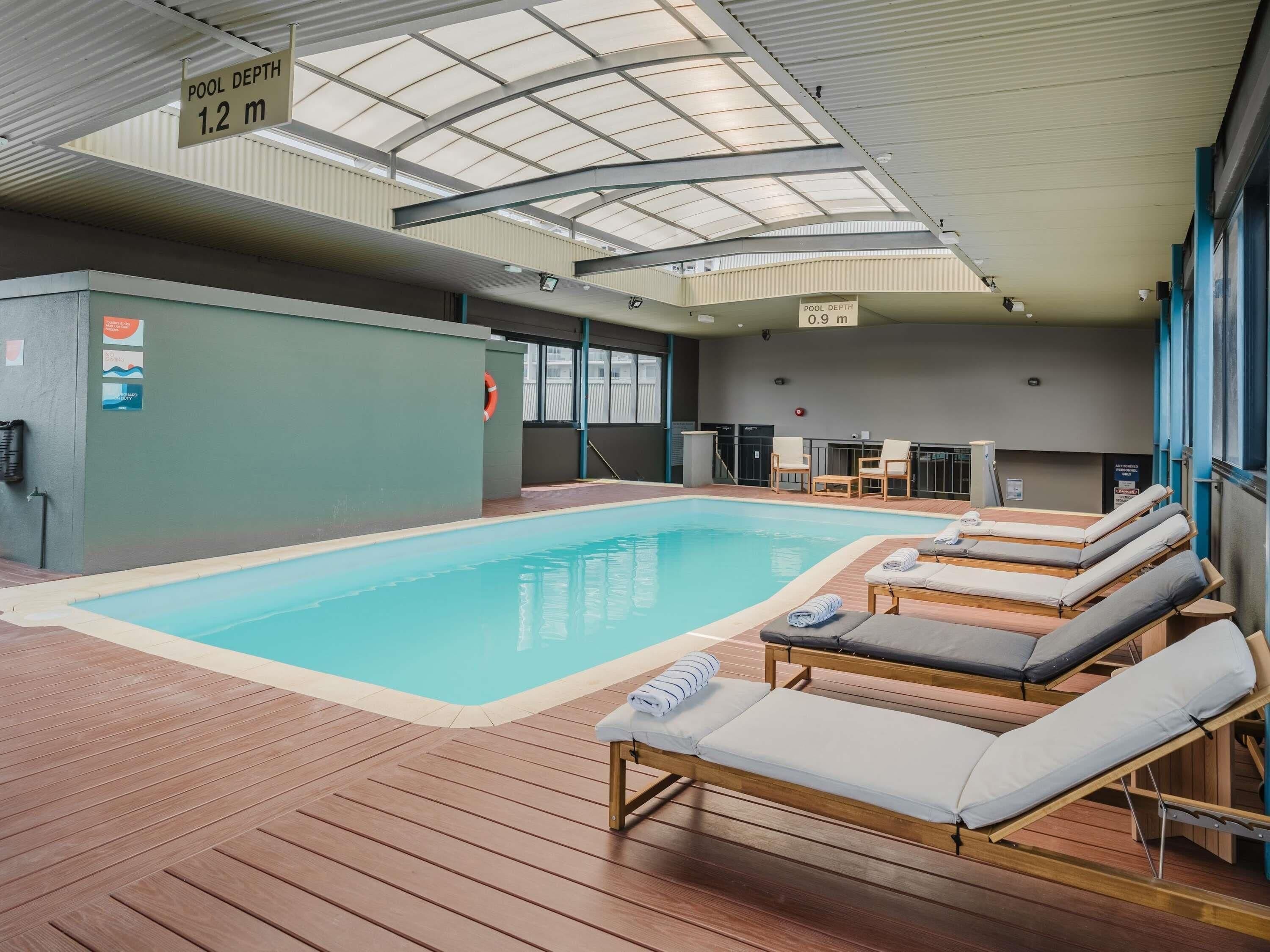 An indoor swimming pool with lounge chairs, a lifebuoy, and signs indicating pool depths.