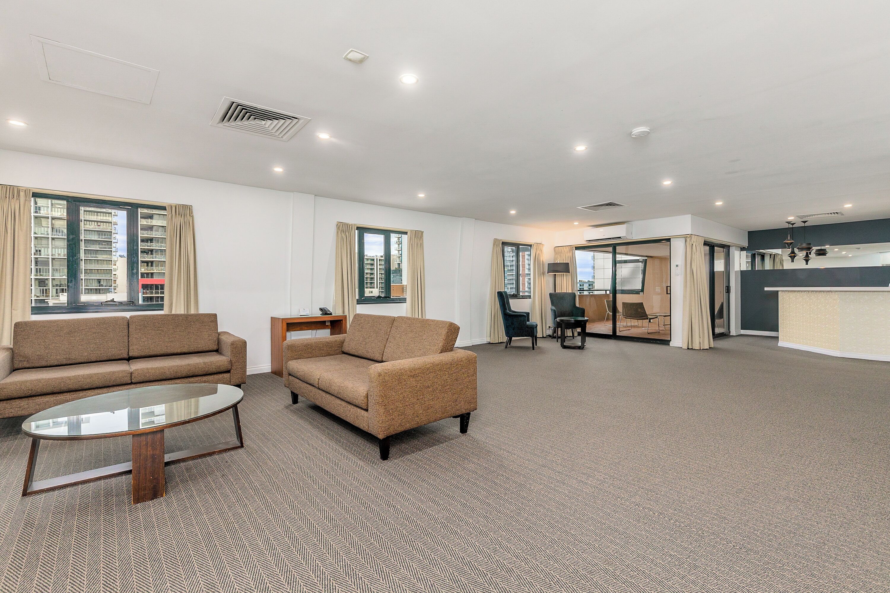 A spacious living area with a sofa, armchair, and coffee table, large windows with curtains, and a view of a high-rise building.