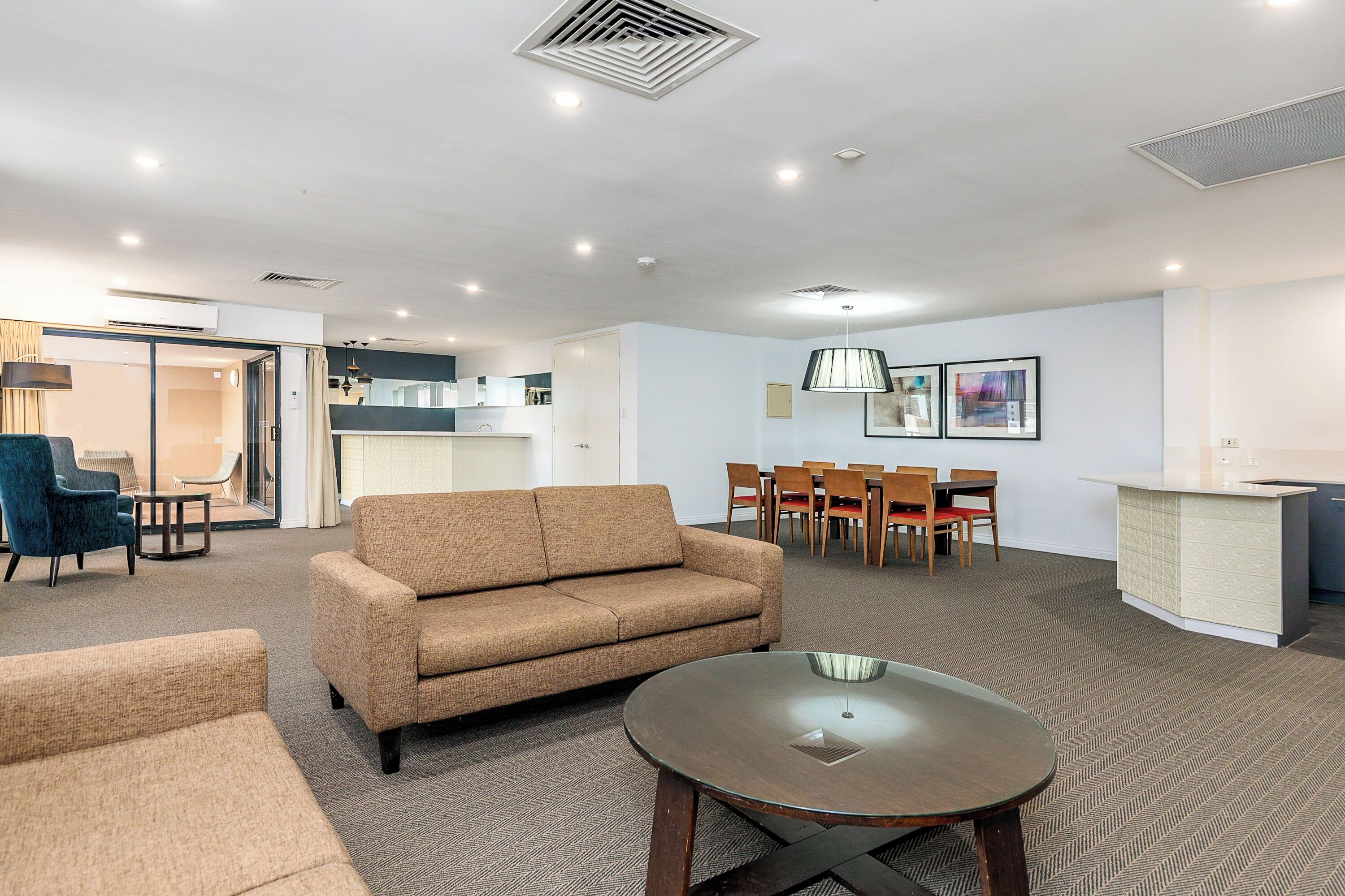 A modern hotel room with a sofa, a round table, a dining area with chairs, a kitchen in the background, and artwork on the wall.