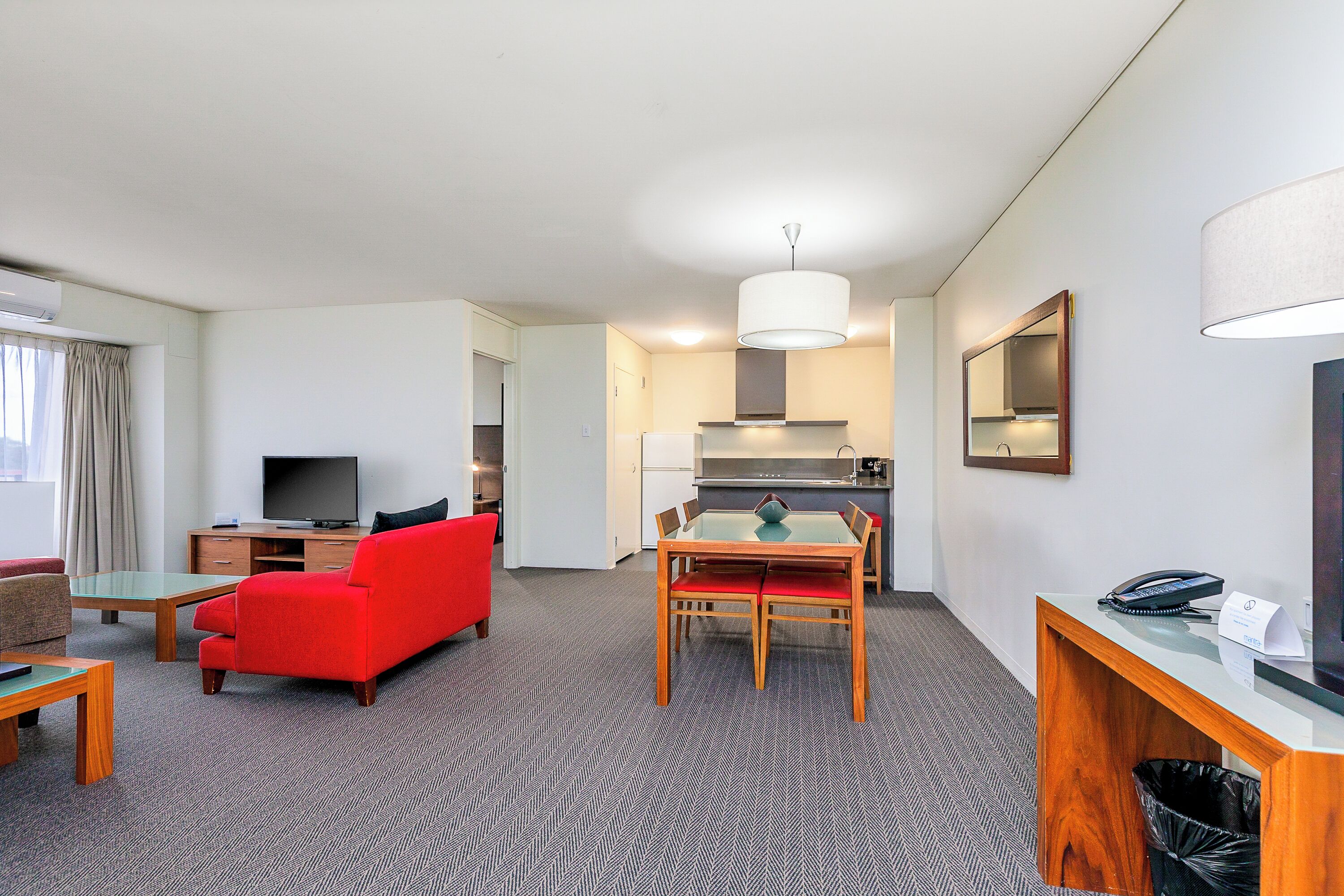 A hotel room with a living area, dining table, red sofa, television, kitchen area, and a desk with a telephone.