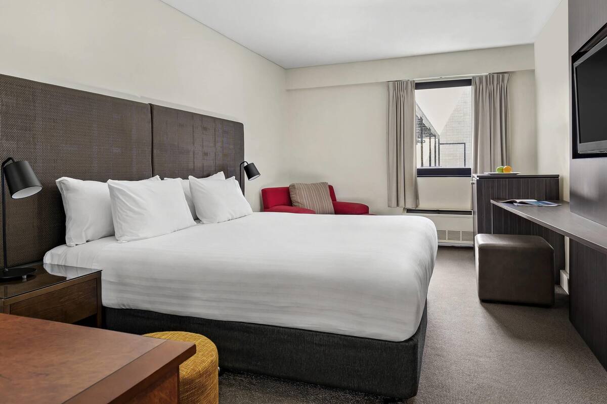 A hotel room with a large bed, a desk, a red chair, a TV, and a window with curtains.