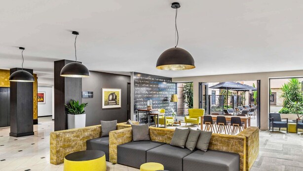 A modern lobby with a sectional sofa, round ottoman, pendant lighting, and a view of the outdoor seating area. - Mantra on Hay Perth (East Perth)