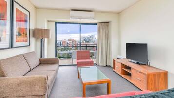 A hotel room with a sofa, armchair, TV, and a balcony with a view of buildings.