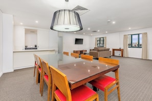 A modern hotel room with a large dining table, wooden chairs, a sofa, a TV, and a window with curtains.