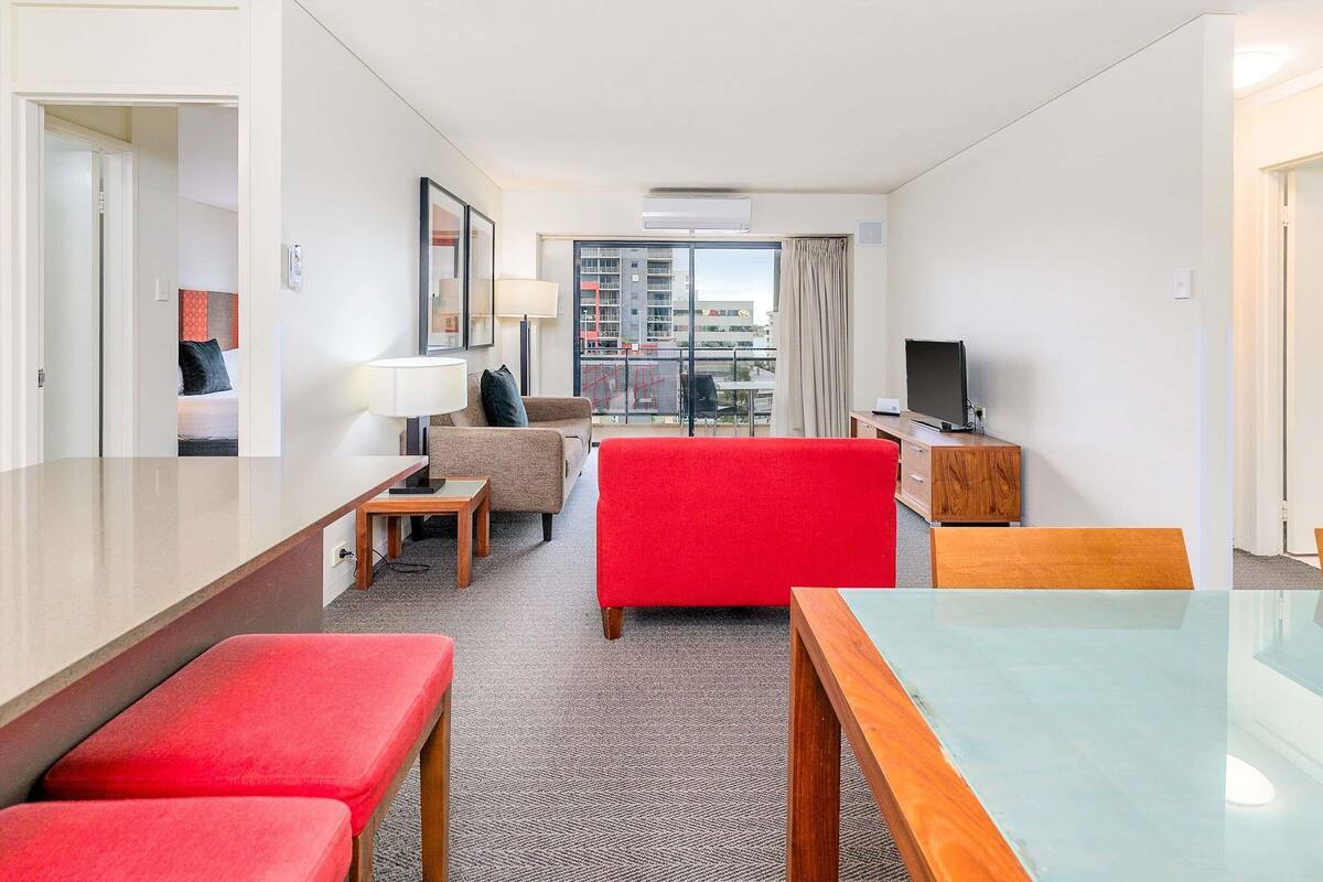 A hotel room with a bed, a red sofa, a wooden desk, a television, and a balcony with a city view.
