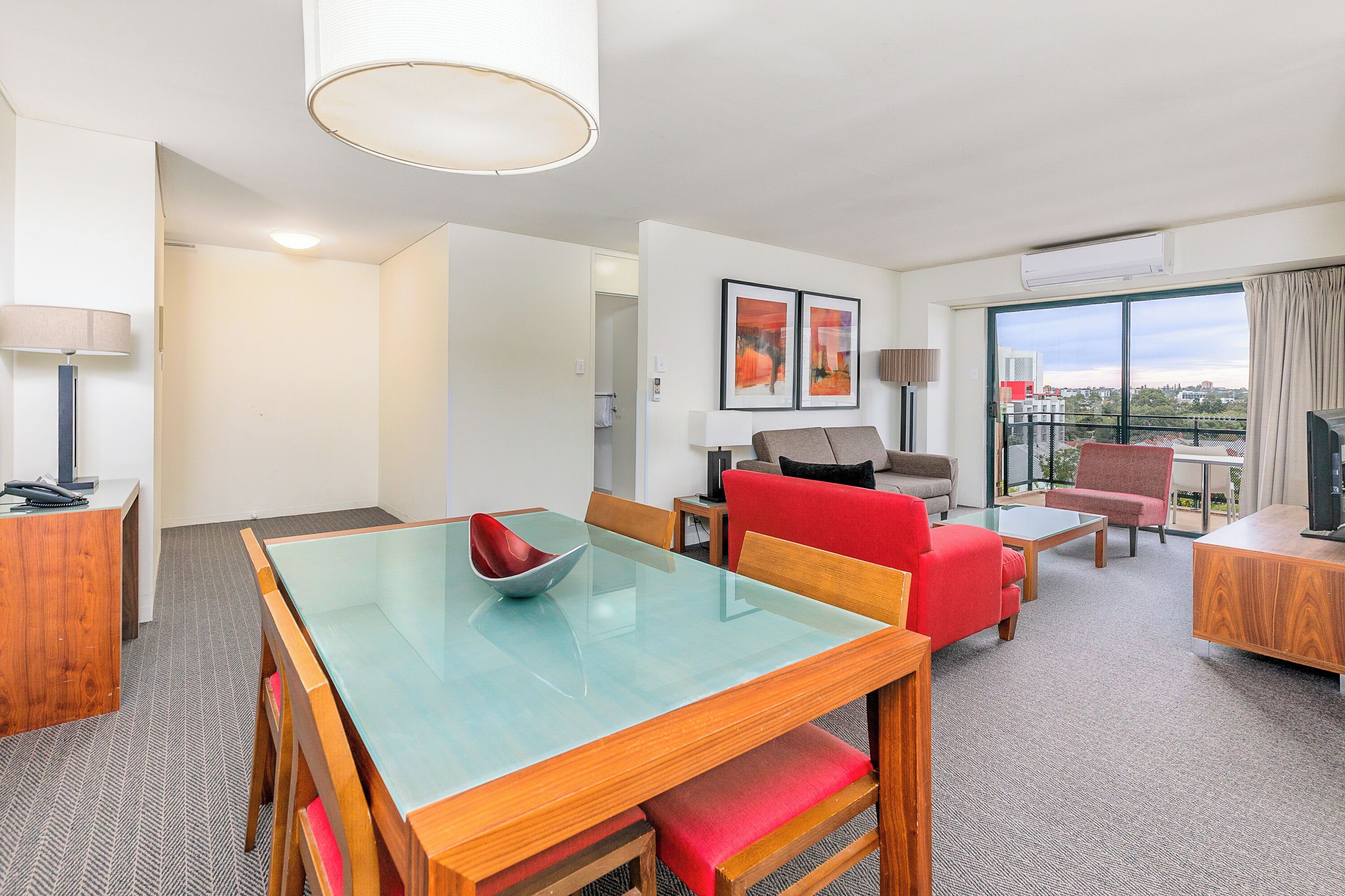 A modern hotel room with a dining area, a living area with red chairs, a TV, and a large window with a city view.