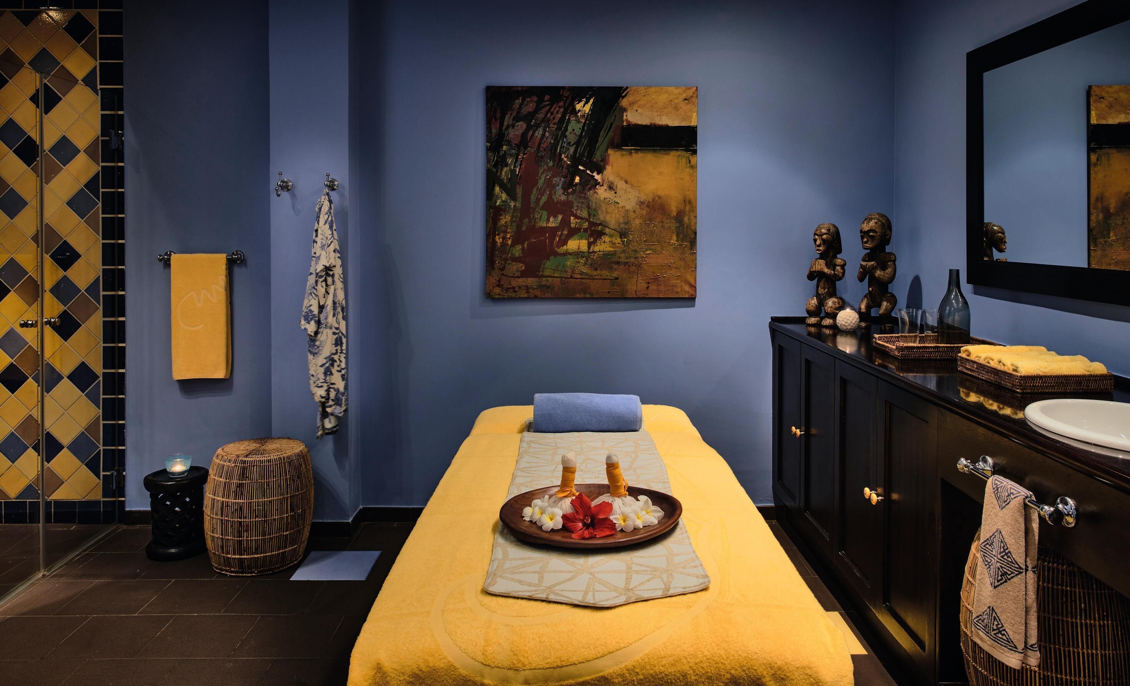 A spa room with a massage table, towels, a painting, and a dark wooden cabinet with a mirror.