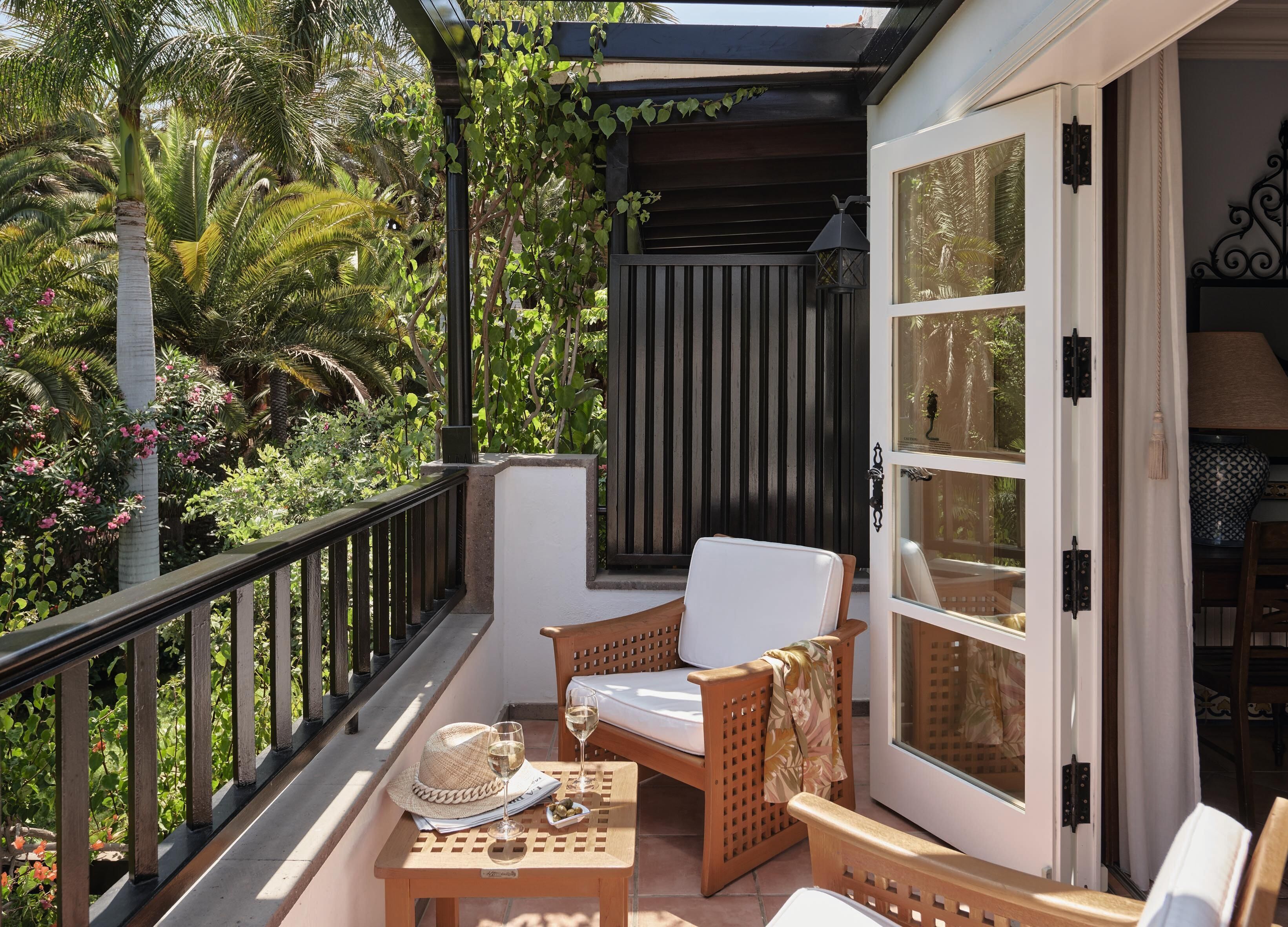 A balcony with a wooden chair, a small table with a hat and glass, and a view of lush greenery and a building with large windows.