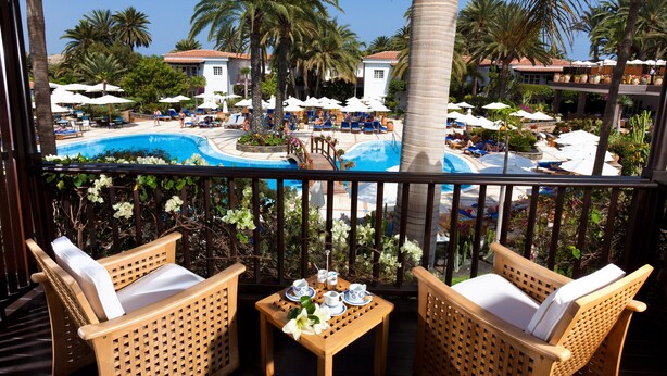 A balcony with wicker chairs and a table set for tea, overlooking a pool area with lounge chairs and umbrellas. - Seaside Grand Hotel Residencia (San Bartolomé de Tirajana)