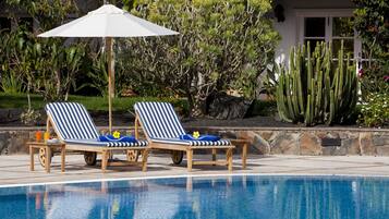 Zona de piscina con dos sillones rayados, una sombrilla blanca y diversos cactus y arbustos al fondo.