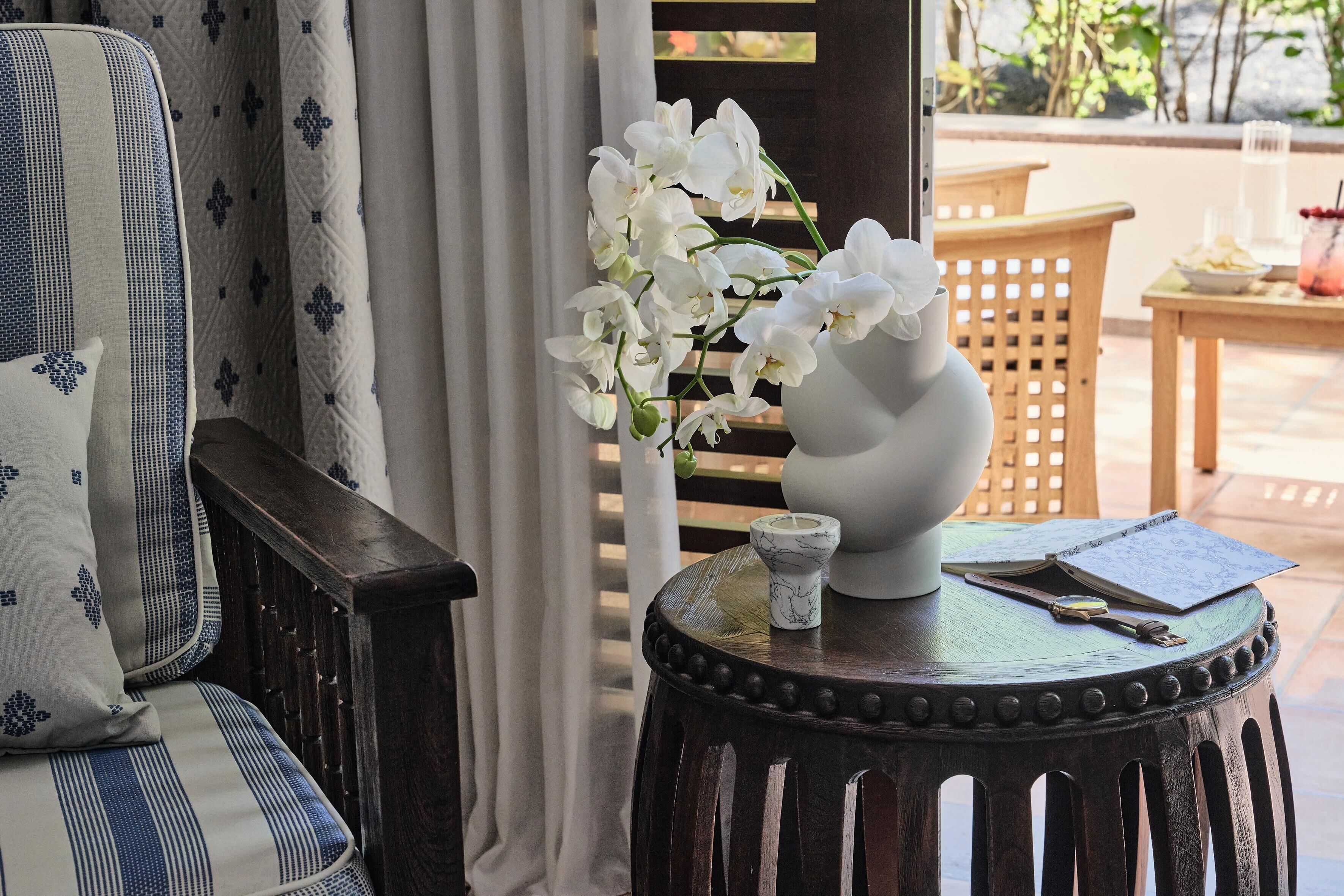 A wooden chair with a cushion, a small round table with a vase of white flowers, and a wooden lattice structure in the background.
