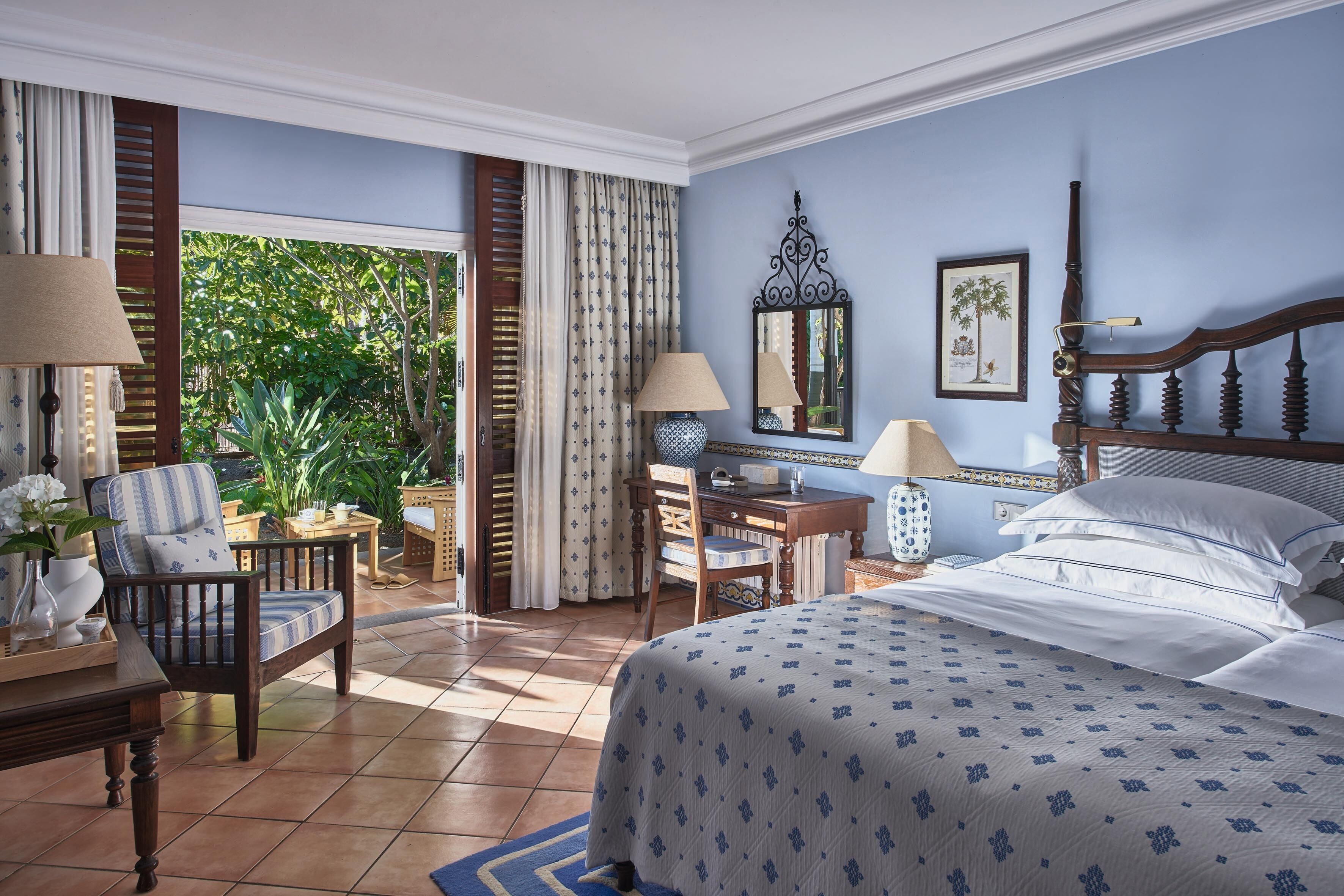 A hotel room with a bed, desk, chair, and a view of greenery outside.