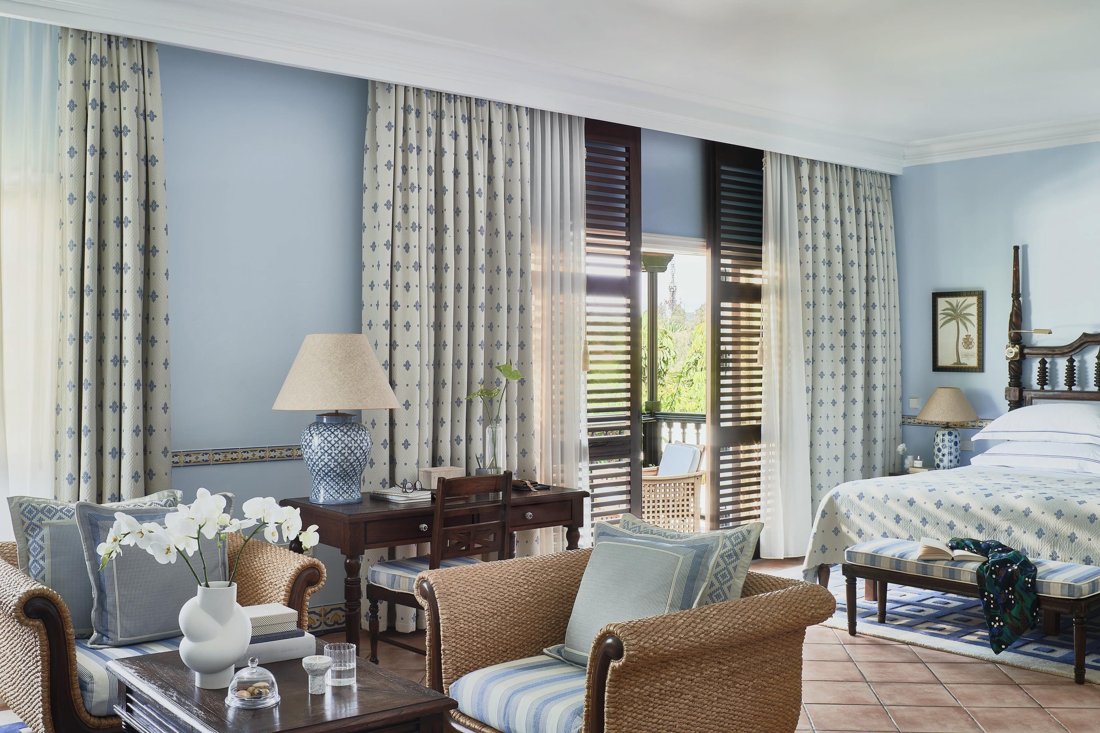 Habitación de hotel con cama, zona de estar con sofá y sillón, mesa de comedor con sillas y ventana con cortinas.