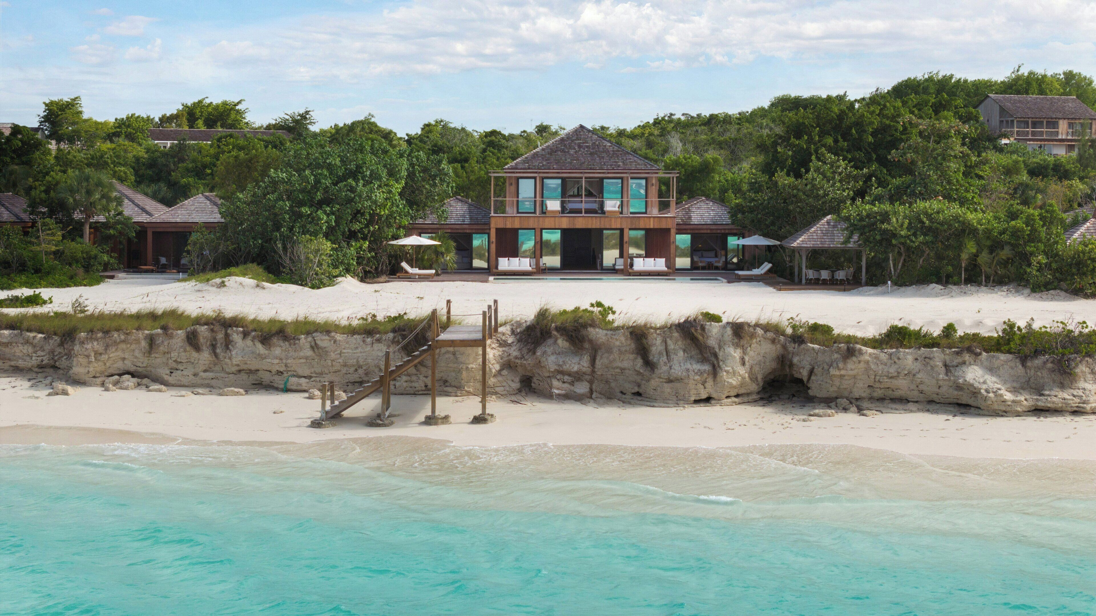 A beachfront property with a large house, a staircase leading to a sandy beach, and a clear view of the ocean.