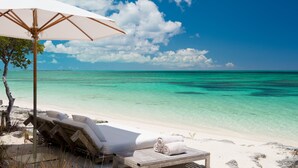 Une plage avec un transat en bois et un parasol, une plage de sable fin et une eau turquoise cristalline.