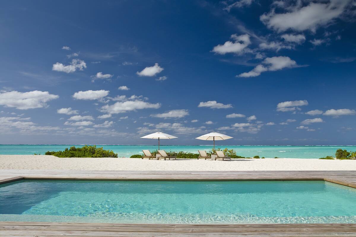 A poolside view with two umbrellas and lounge chairs on a wooden deck, overlooking a beach and ocean.