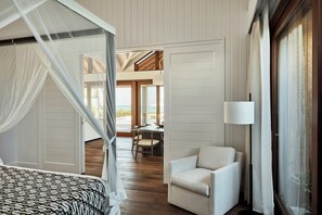 A bedroom with a four-poster bed, a sitting area with a chair, and a view of the beach through an open door.