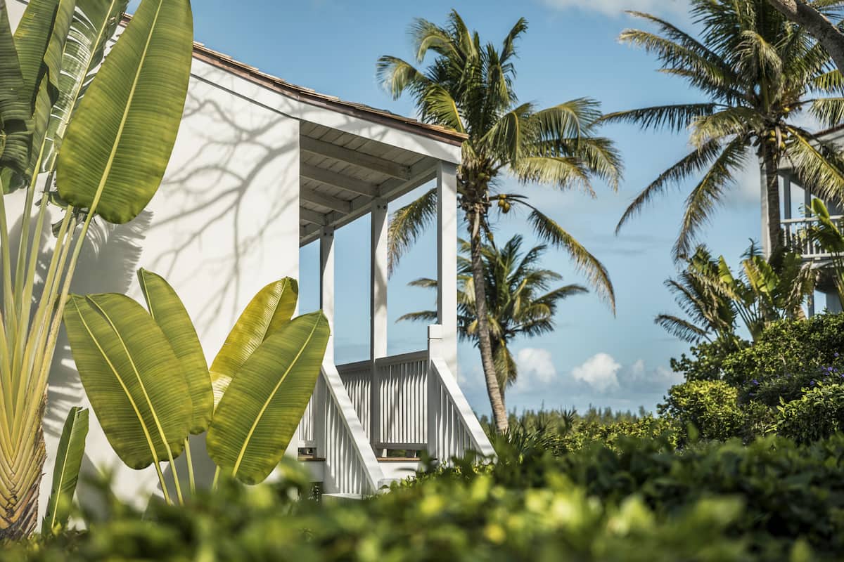 A white building with a covered porch, surrounded by tropical vegetation and palm trees.