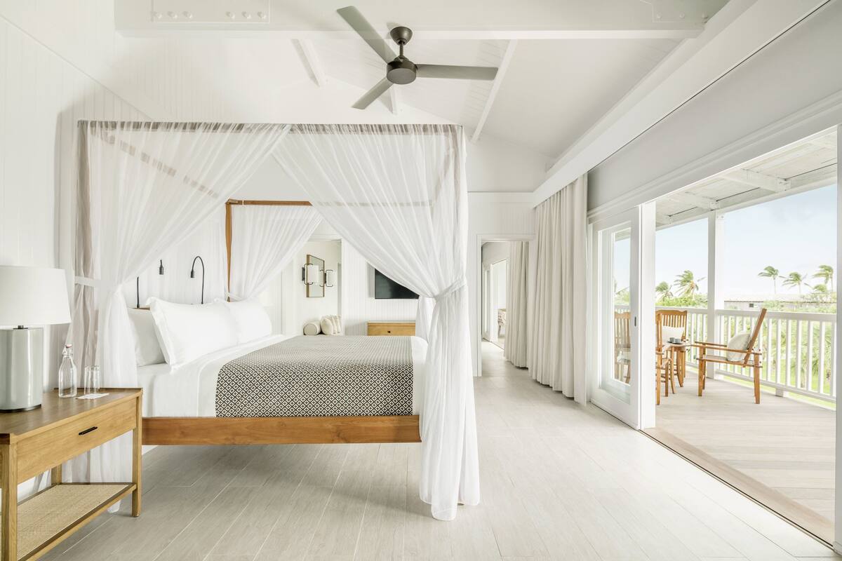A bedroom with a canopy bed, a ceiling fan, a wooden nightstand, and a view of the balcony.