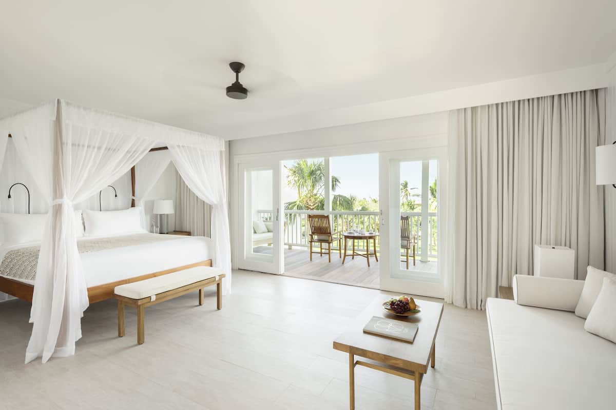 A bedroom with a four-poster bed, a bench, a bedside table, a sofa, and a view of the outdoor balcony.