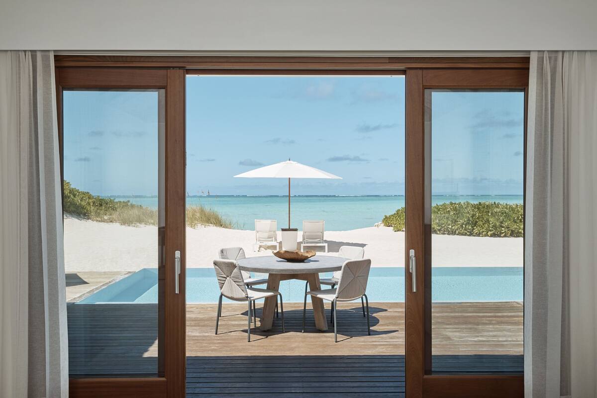 A view from a sliding glass door to a deck with a round table and chairs, overlooking a beach and ocean.