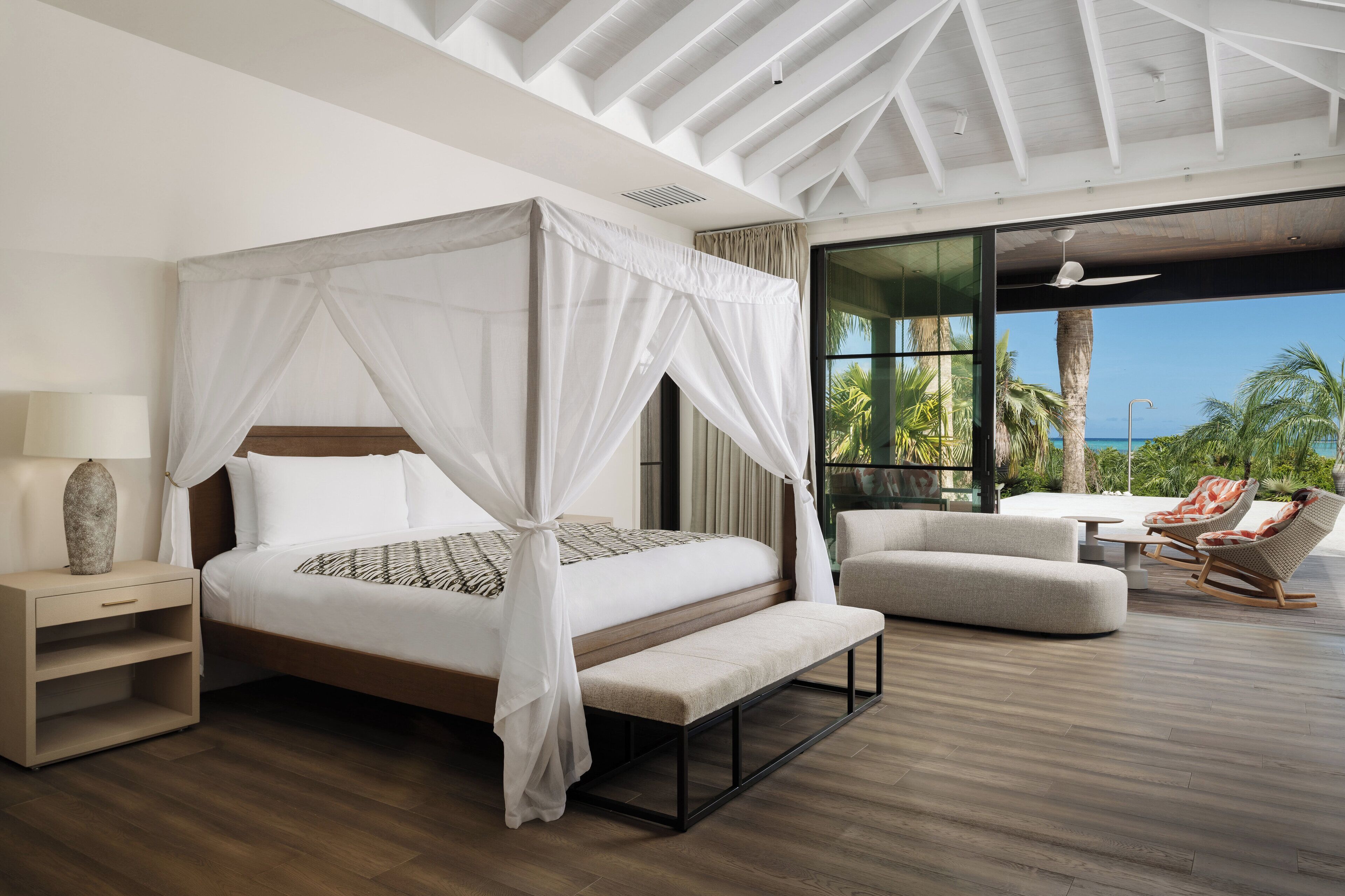 Ein Schlafzimmer mit einem Himmelbett, einer Bank, einem Nachttisch und Meerblick durch große Fenster.