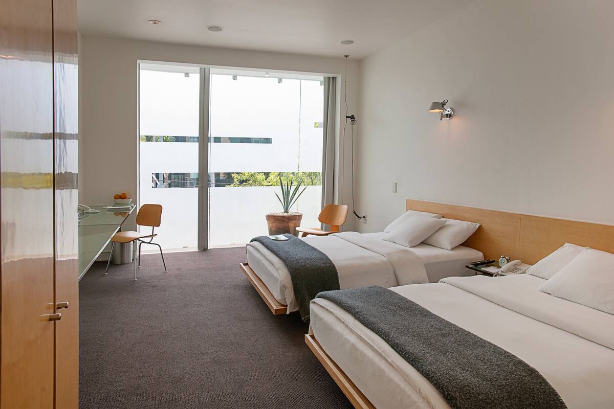 A hotel room with two beds, a desk, a chair, and a view of the outdoors.