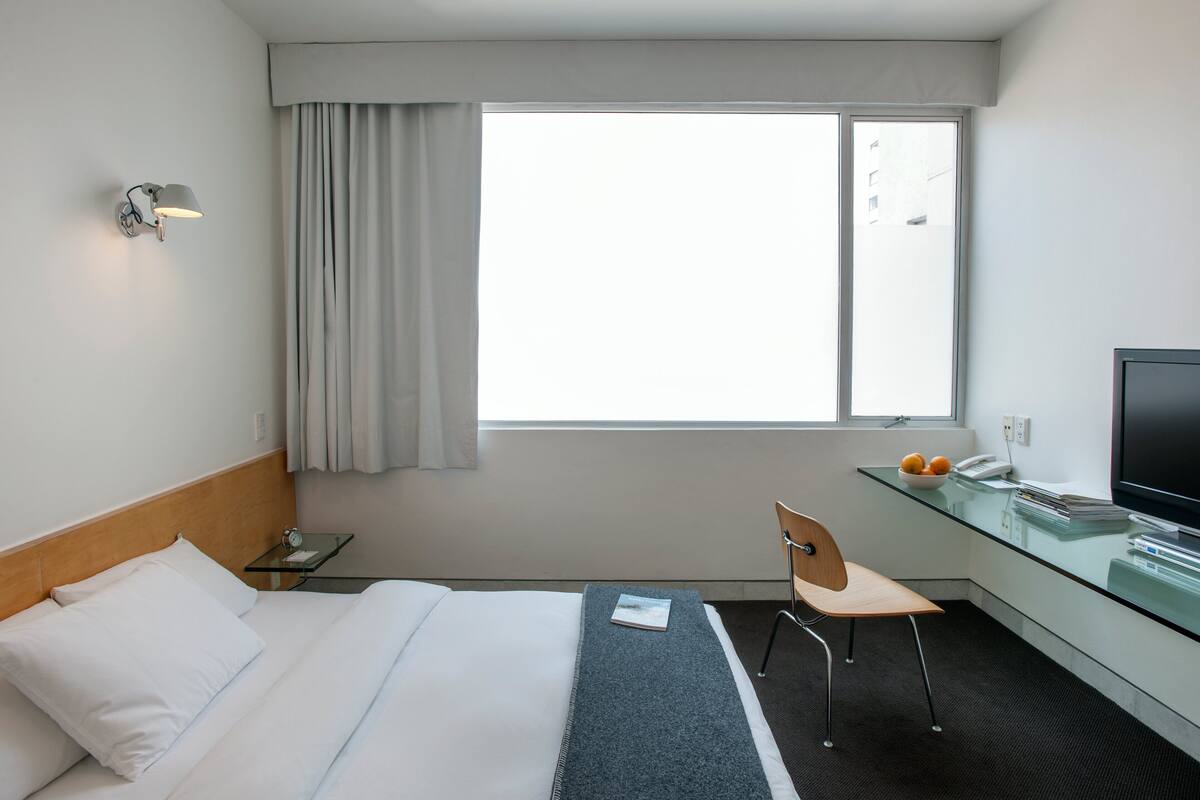 A hotel room with a bed, a desk with a chair, a TV, and a window with curtains.
