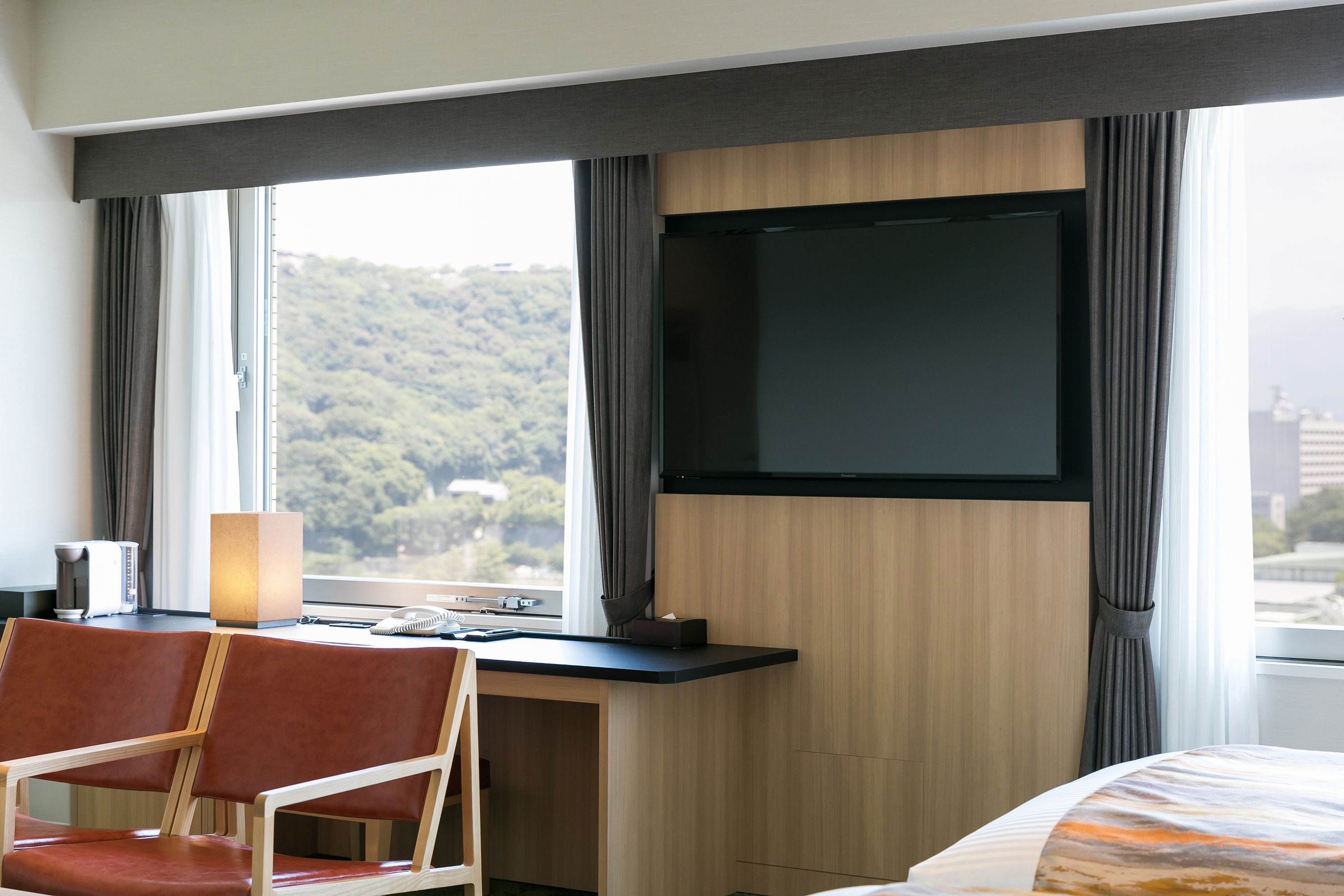 A hotel room with a desk, a chair, a bed, a TV mounted on the wall, and a window with a view of a forested area.