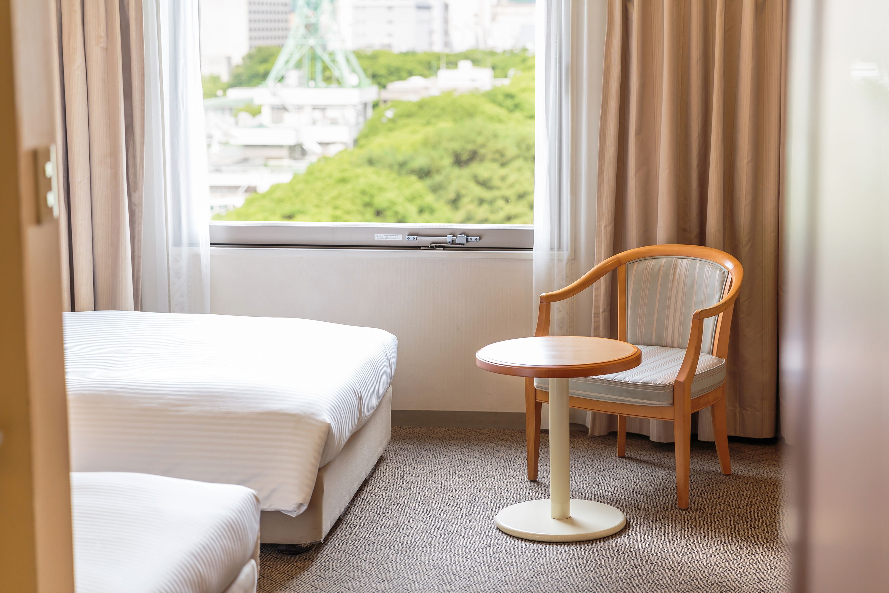 A hotel room with a bed, a chair, and a window with a view of greenery and buildings.
