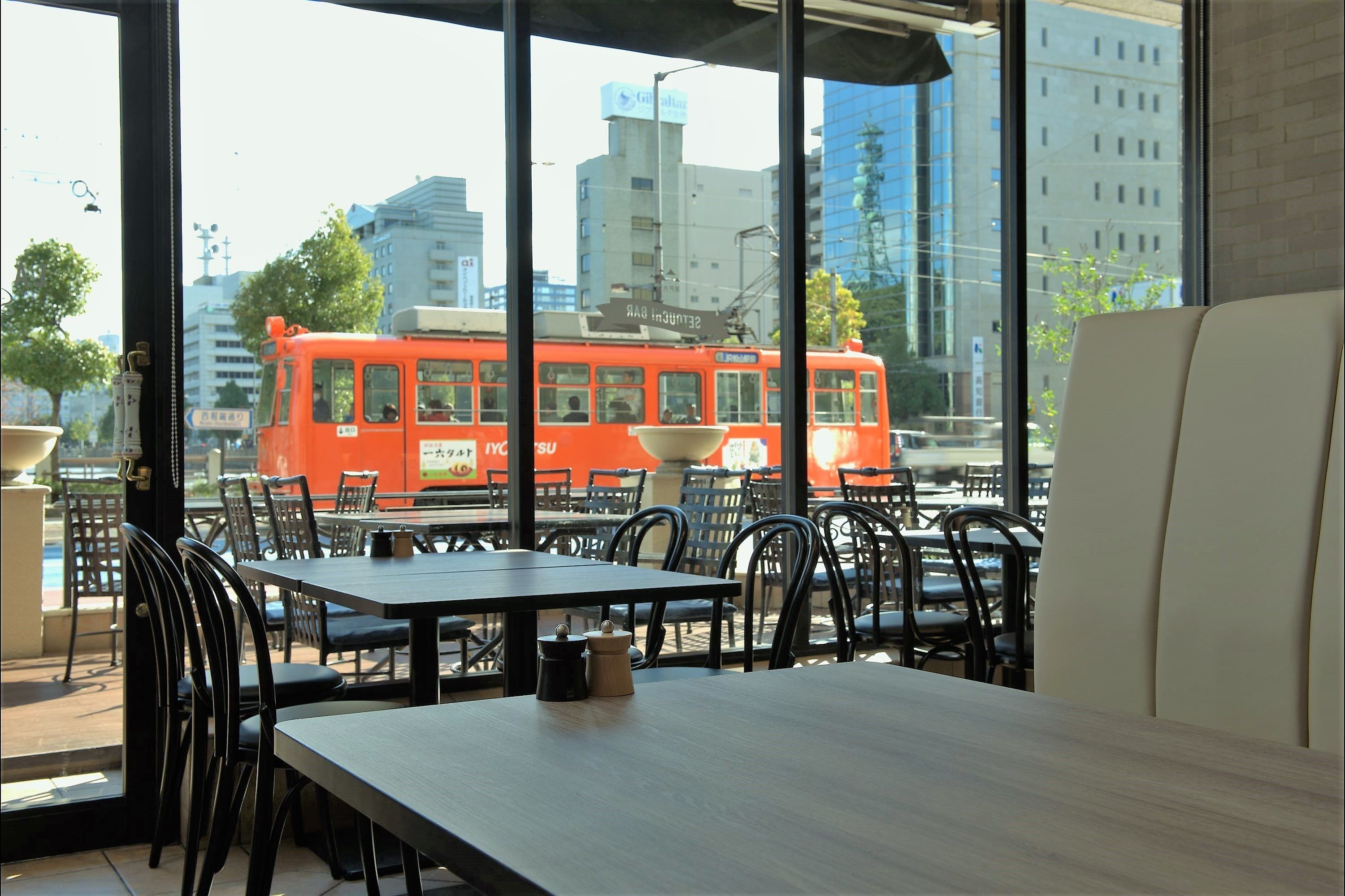 A restaurant with outdoor seating, tables, chairs, and a view of a red tram in the background.