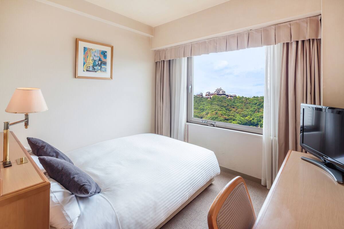 Une chambre d’hôtel avec un lit, un bureau avec une télévision, une fenêtre avec des rideaux, un tableau au mur et une lampe.