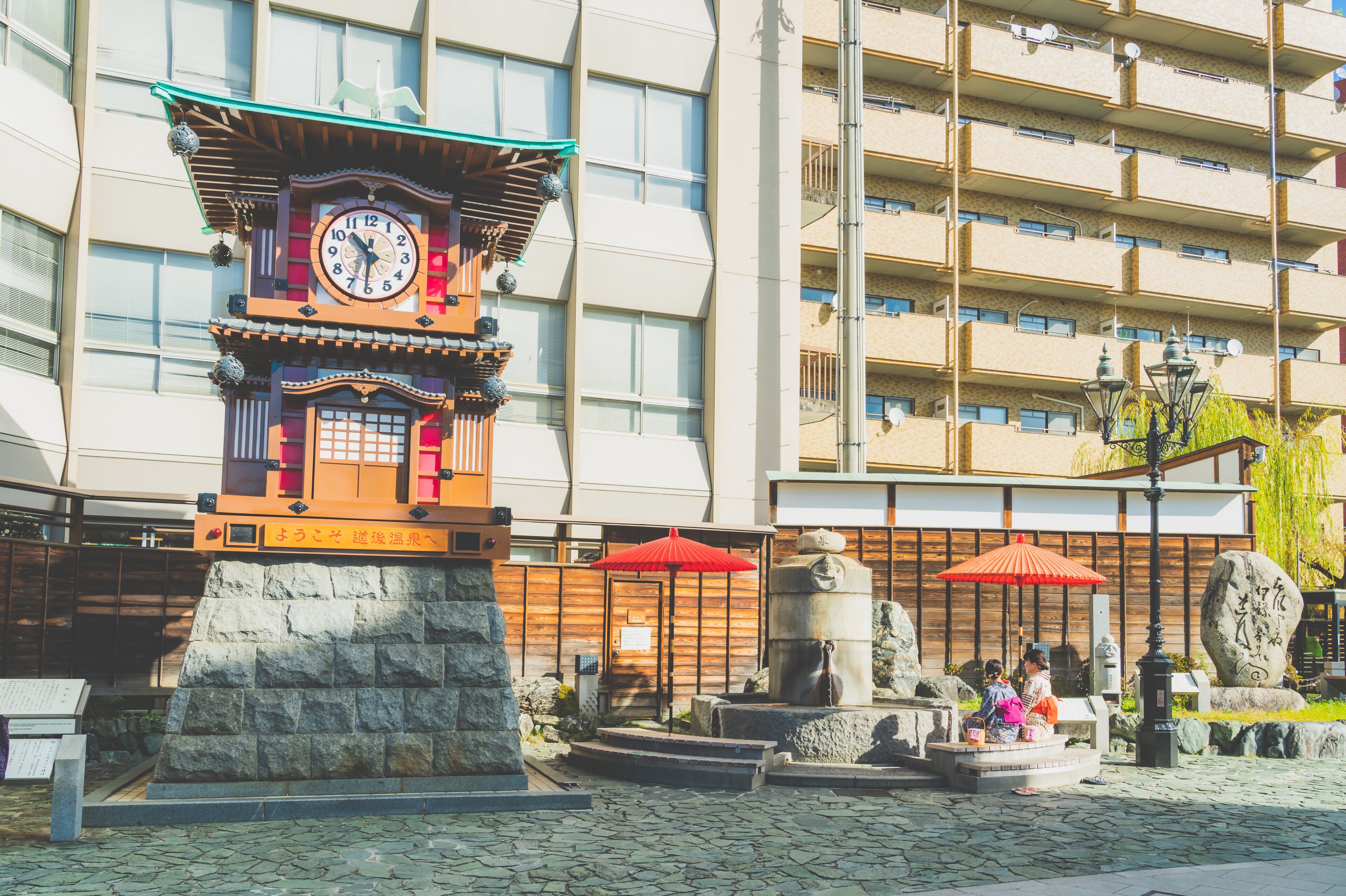 Une tour de l’horloge de style japonais traditionnel, avec une base en pierre, entourée d’un sentier pavé et de parasols rouges.