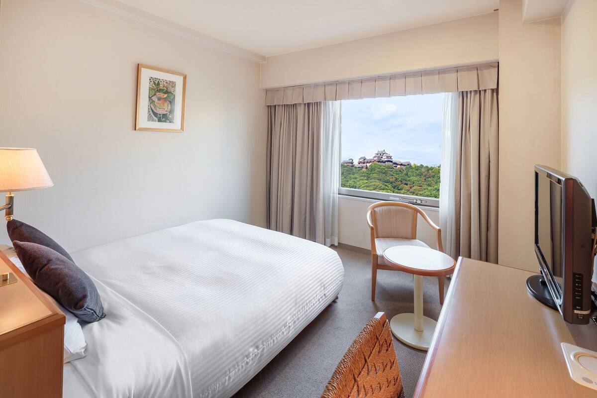 Une chambre d’hôtel comprenant un lit, un bureau avec un ordinateur, une chaise, une télévision et une fenêtre avec des rideaux.