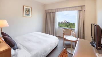 A hotel room with a bed, a desk with a computer, a chair, a TV, and a window with curtains.