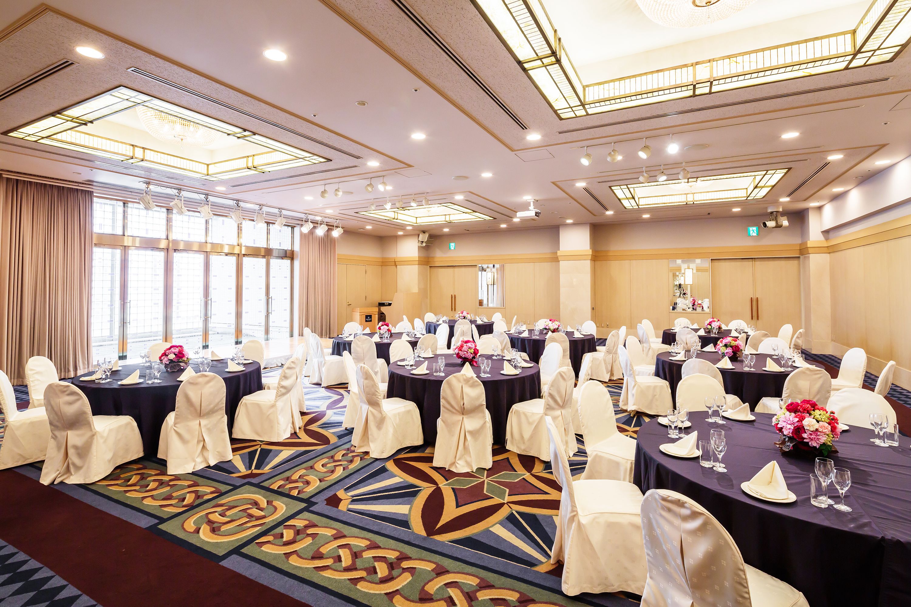 A large conference room with multiple round tables, each set with white chairs and tablecloths, and floral centerpieces.