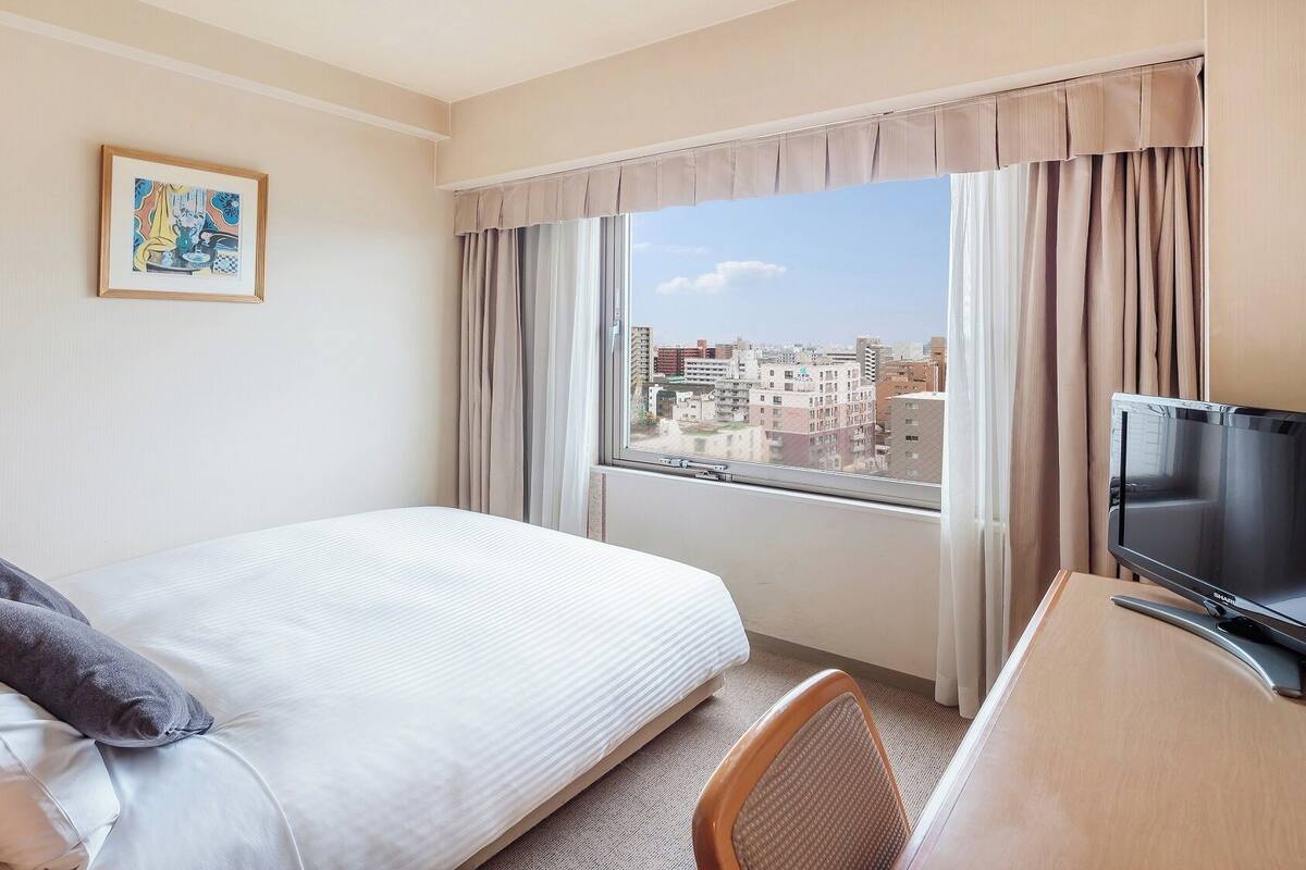 Une chambre d’hôtel avec un lit, un bureau doté d’une lampe, une télévision, une fenêtre avec des rideaux et un tableau encadré au mur.