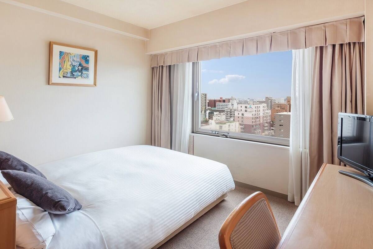 Une chambre d’hôtel avec un lit, un bureau avec une télévision, une fenêtre avec des rideaux et un tableau au mur.