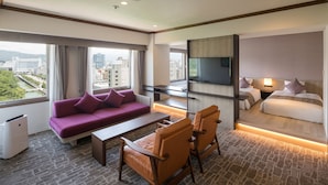 A hotel room with a sofa, two armchairs, a bed, a TV, and a city view.