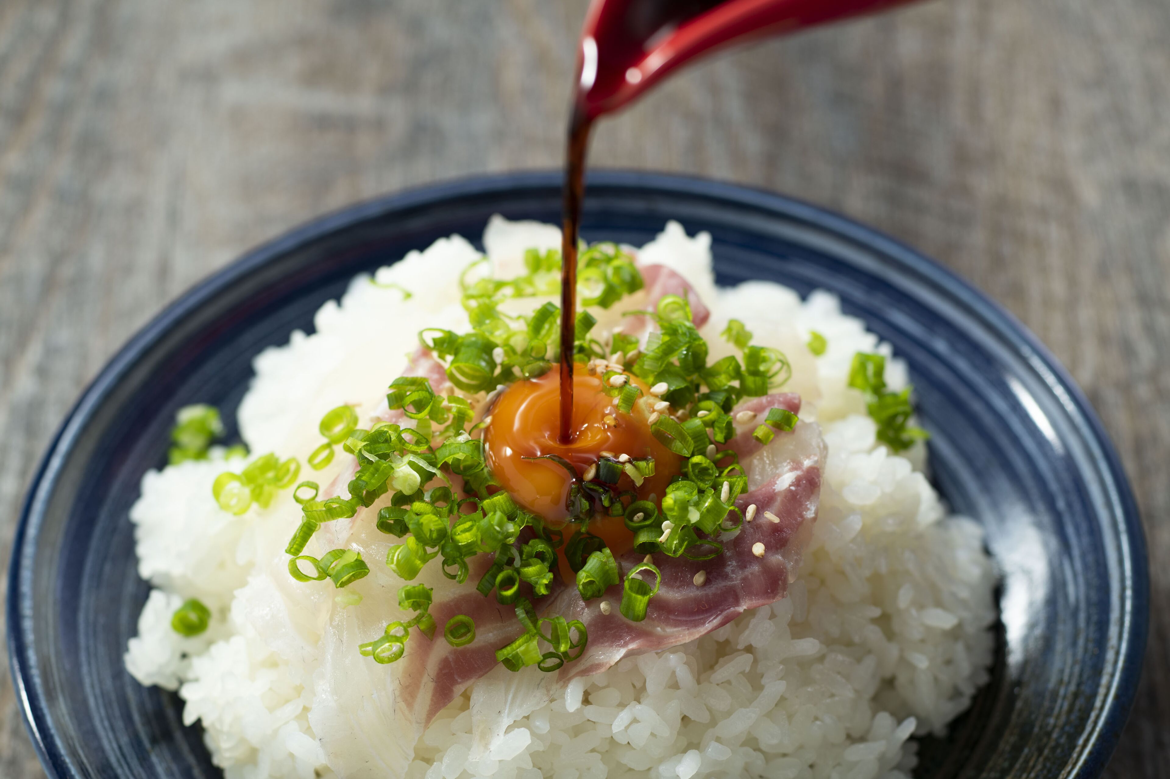 A bowl of rice topped with green onions, a slice of meat, and a sauce being poured over it.