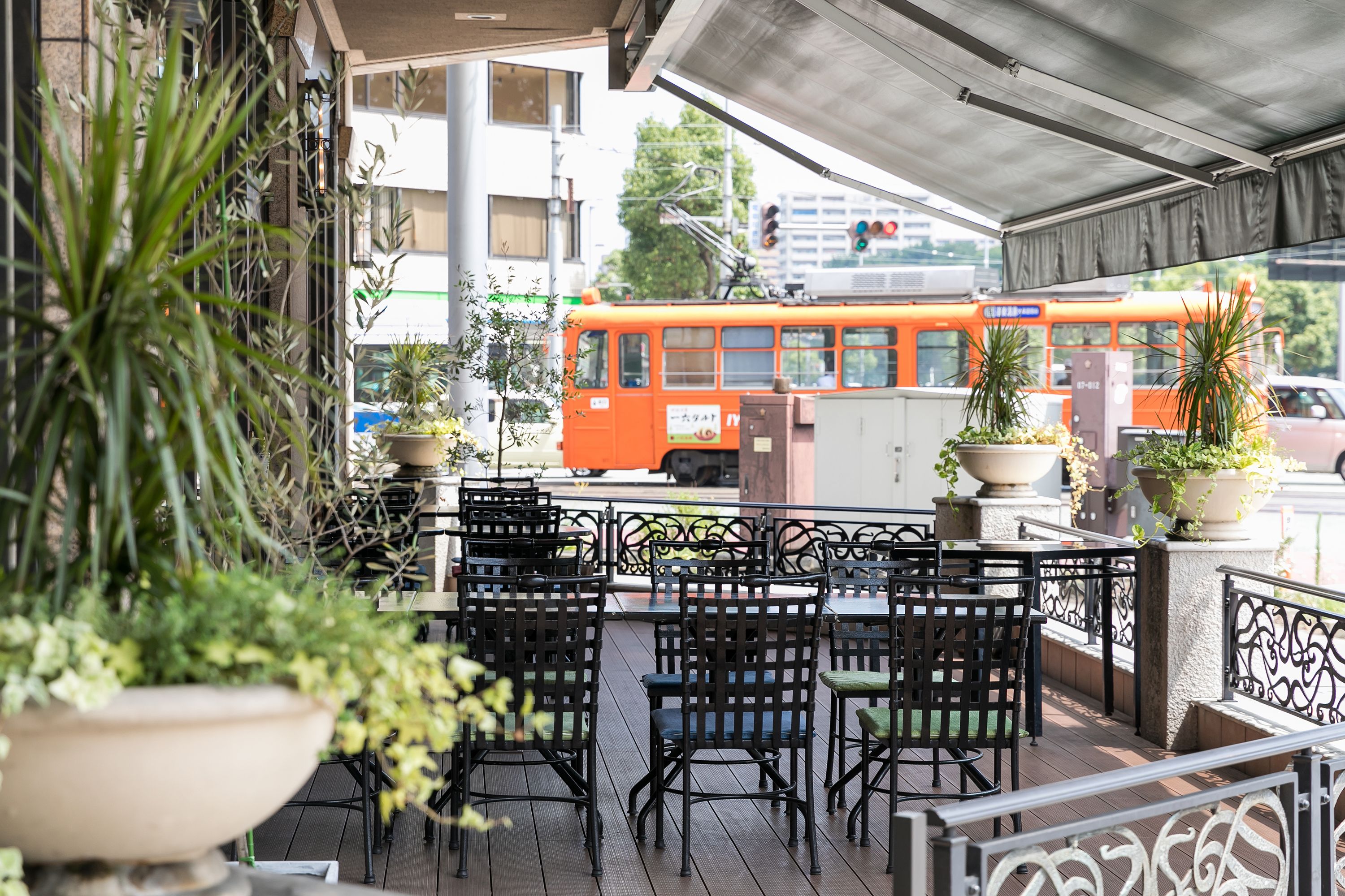 An outdoor seating area with tables and chairs, potted plants, and a view of a city street with a bus.