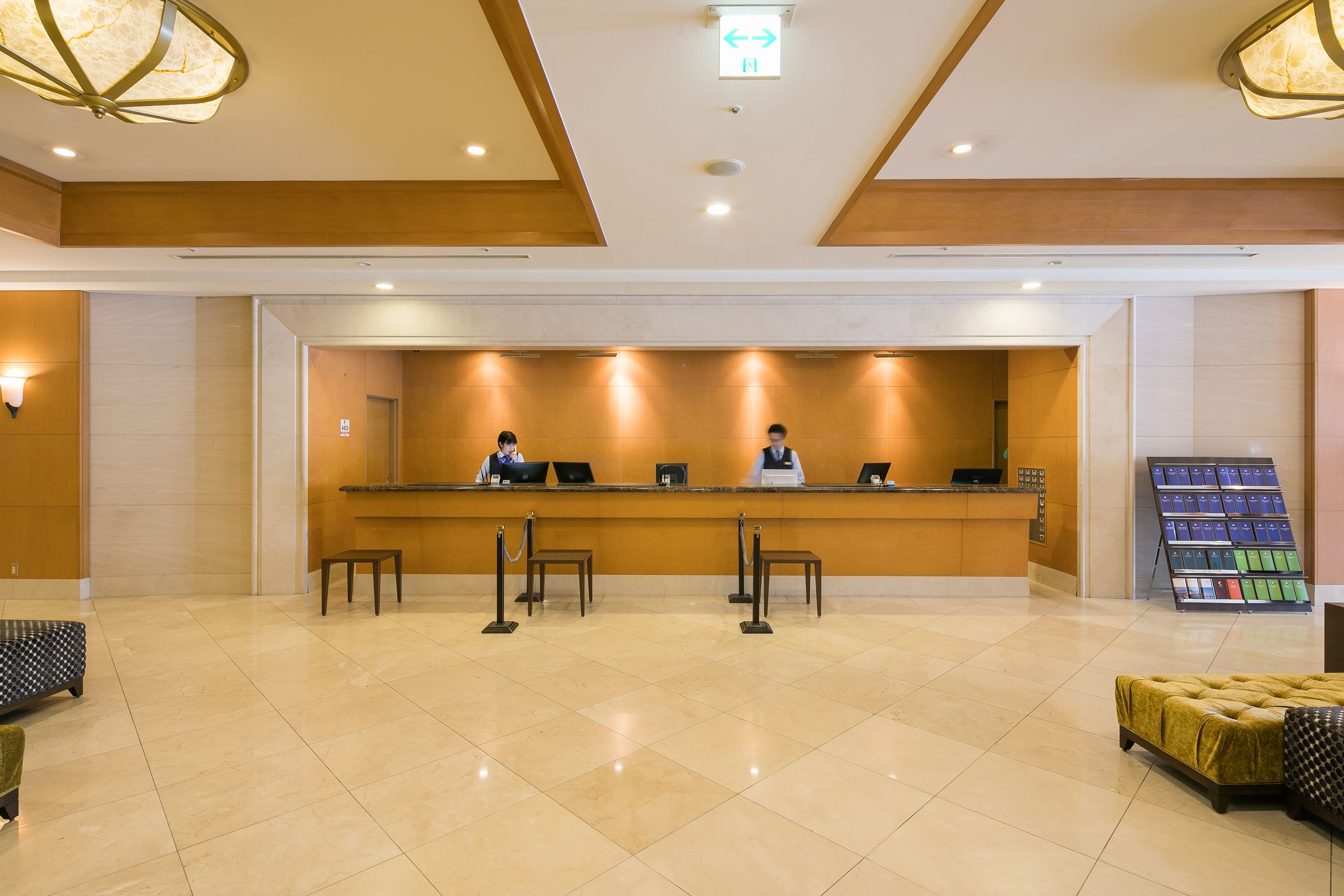 Zona de recepción con piso de mármol, mostrador de madera, dos recepcionistas y una exhibición de folletos.