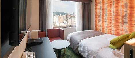 Habitación de hotel con cama, escritorio, silla, mesita, teléfono y ventana con vistas a edificios y una colina.