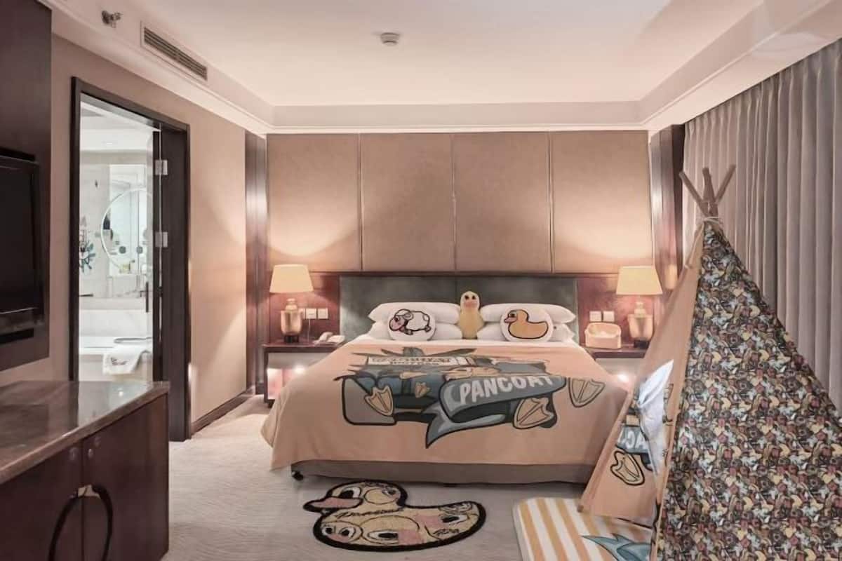 Un dormitorio con cama, lámparas de noche, mesita de noche, televisor, y un baño visible a través de una puerta abierta, además de una alfombra estampada.