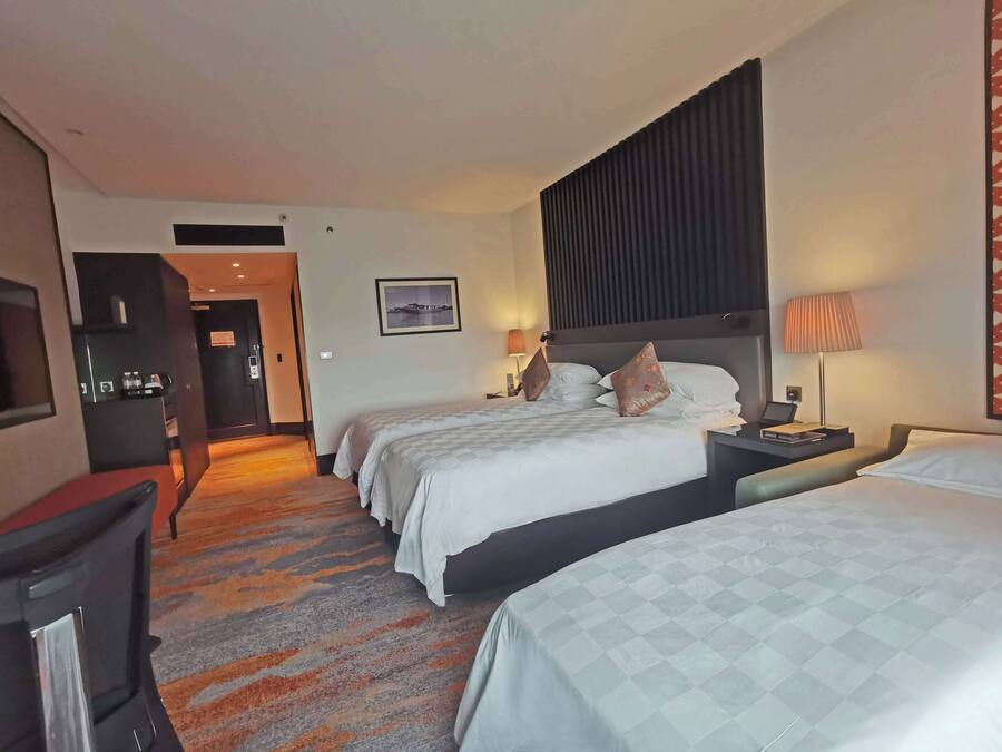 Habitación de hotel con dos camas, un escritorio, una silla, un televisor y una puerta que da a un pasillo.