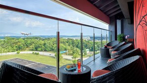 Ein Balkon mit Korbmöbeln, einem Getränk auf dem Tisch und einem Flugzeug in der Ferne.